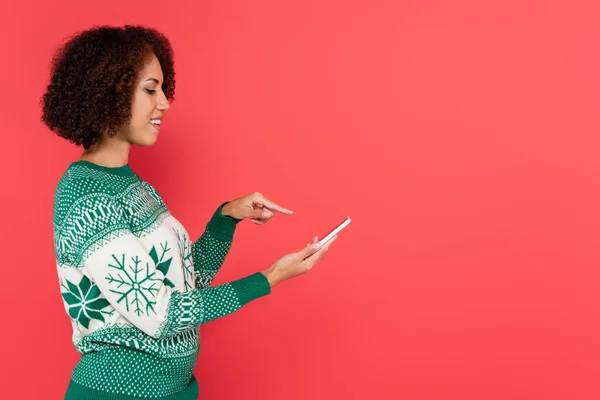 Vue latérale de la femme brune afro-américaine en pull chaud pointant du doigt le smartphone isolé sur rouge — Photo de stock