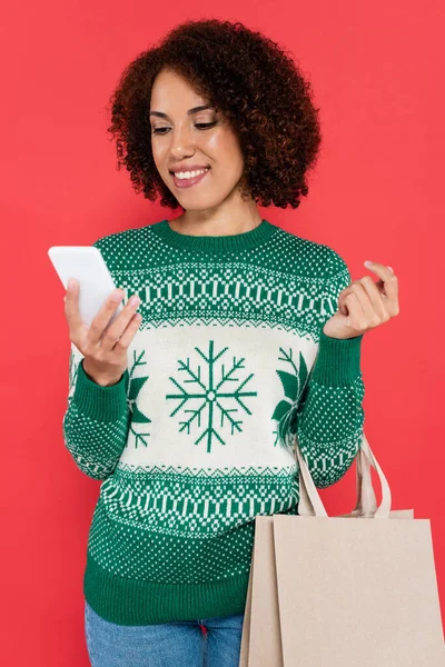 Sonriente mujer afroamericana en suéter con patrón de invierno de pie con bolsas de compras y teléfono inteligente aislado en rojo - foto de stock