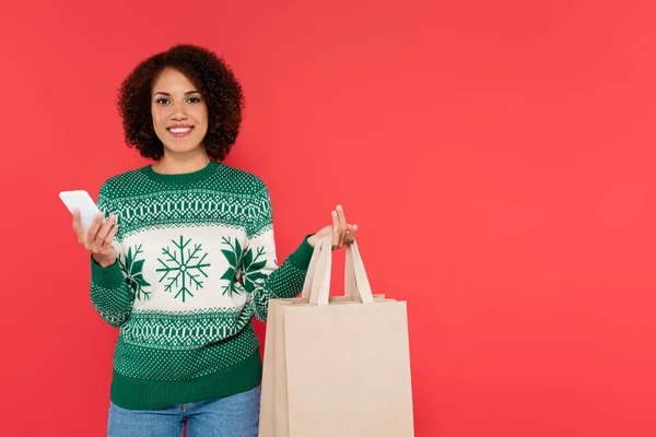 Alegre mujer afroamericana en suéter caliente sosteniendo bolsas de compras y teléfono inteligente aislado en rojo - foto de stock