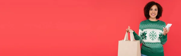 Jovem afro-americana com sacos de compras e telefone celular sorrindo para câmera isolada em vermelho, banner — Fotografia de Stock