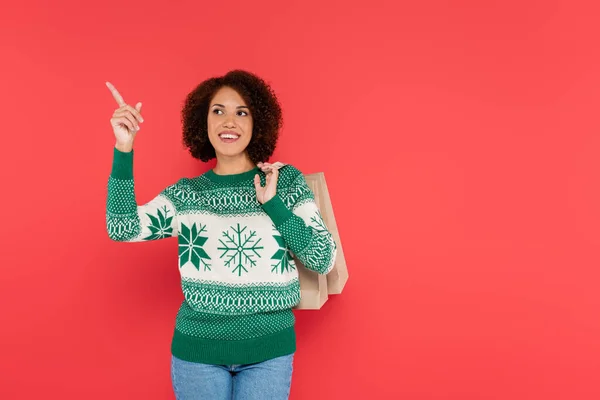 Smiling african american woman in white sweater with green ornament pointing with finger and looking away isolated on red — Stock Photo