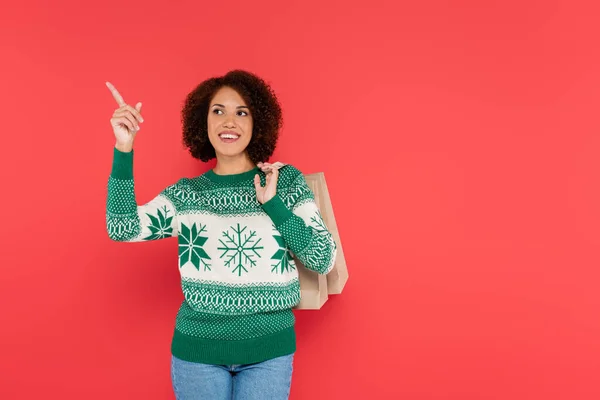 Donna afroamericana sorridente in maglione bianco con ornamento verde che punta con il dito e distoglie lo sguardo isolato sul rosso — Foto stock