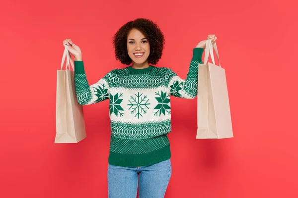Pleased african american woman in white sweater with green pattern showing shopping bags isolated on red — Stock Photo