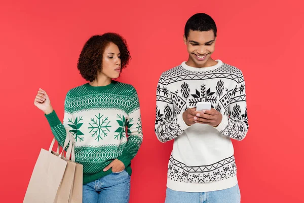 Mulher americana africana curioso com sacos de compras olhando para namorado conversando no smartphone isolado no vermelho — Fotografia de Stock