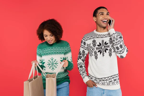 Mujer afroamericana mirando en bolsas de compras cerca de hombre alegre hablando en el teléfono celular aislado en rojo - foto de stock