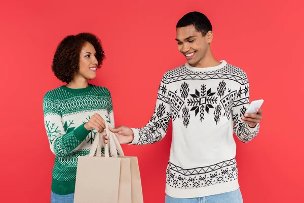 Sonriente mujer afroamericana sosteniendo bolsas de compras cerca de hombre con teléfono inteligente aislado en rojo - foto de stock