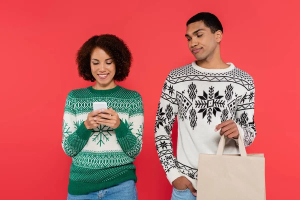 Hombre afroamericano disgustado con bolsa de compras mirando a la novia charlando en el teléfono inteligente aislado en rojo - foto de stock