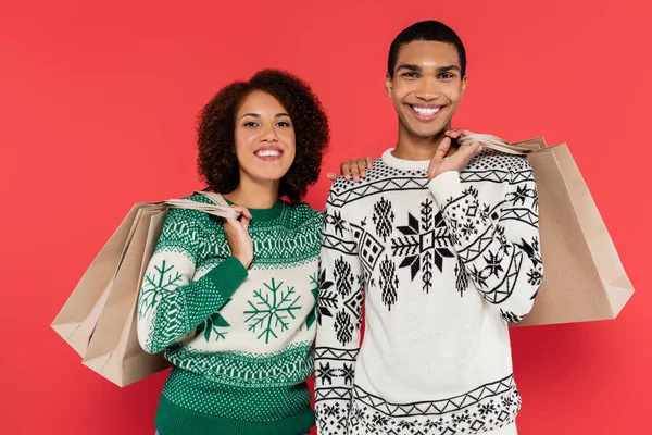 Alegre pareja afroamericana en suéteres de invierno sosteniendo bolsas de compras y mirando a la cámara aislada en rojo - foto de stock