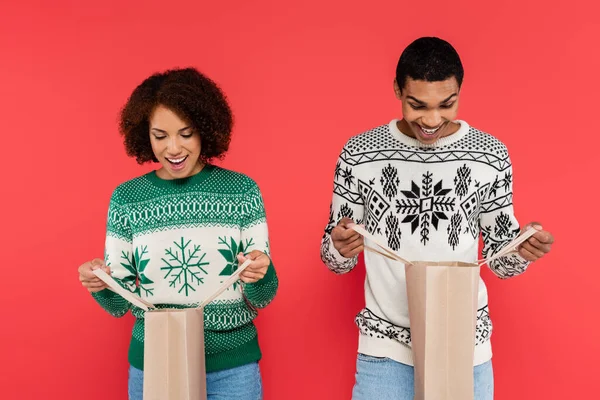 Asombrada pareja afroamericana en suéteres de invierno mirando en bolsas de compras aisladas en rojo - foto de stock