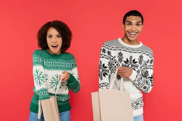 Excitada pareja afroamericana en suéteres calientes revisando bolsas de compras aisladas en rojo - foto de stock