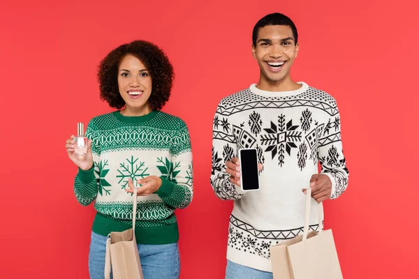 Joyeux couple afro-américain tenant des sacs à provisions et smartphone avec parfum isolé sur rouge — Photo de stock