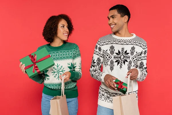 Coppia afro-americana con borse della spesa e scatole regalo verdi sorridenti l'un l'altro isolati su rosso — Foto stock