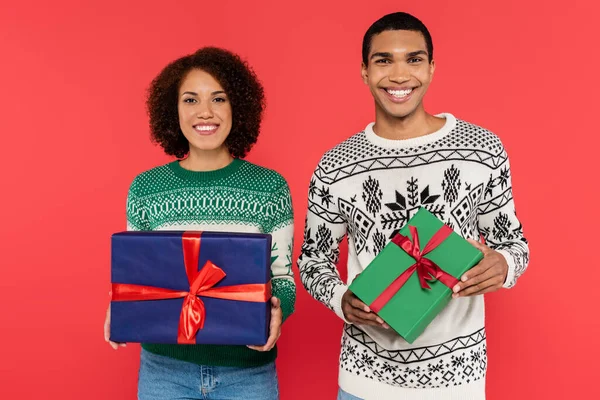 Alegre pareja afroamericana con cajas de regalo verdes y azules mirando a la cámara aislada en rojo - foto de stock