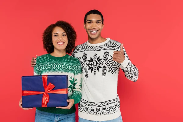 Souriant homme afro-américain montrant pouce vers le haut près de petite amie heureuse avec cadeau de Noël isolé sur rouge — Photo de stock