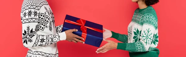 Cropped view of african american couple in warm sweaters holding blue gift box isolated on red, banner — Stock Photo