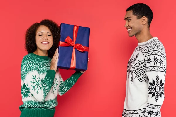 Pleased african american woman with closed eyes holding blue gift box near smiling boyfriend isolated on red — Stock Photo