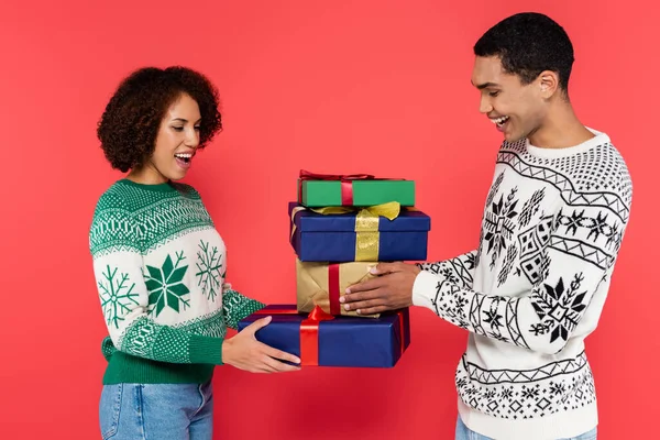 Зачаровані парою афроамериканців у теплих светрах з зимовим орнаментом, що тримає новорічні подарунки ізольованими на червоному — стокове фото