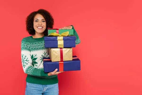 Pleased african american woman in winter sweater holding stack of gift boxes and looking at camera isolated on red — Stock Photo