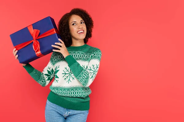 Sorrindo e mulher americana africana curiosa em suéter de inverno balançando caixa de presente azul isolado no vermelho — Fotografia de Stock