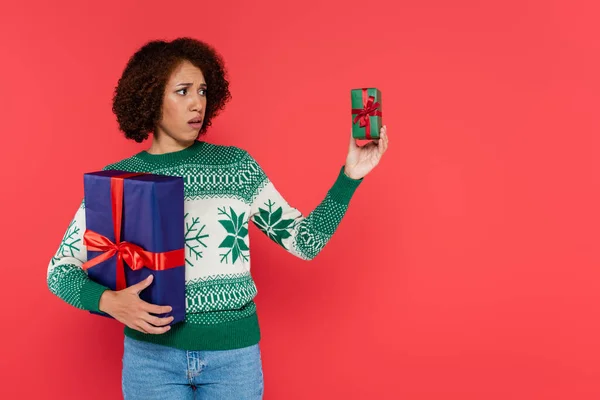 Displeased african american woman with big gift box looking at small present isolated on red — Stock Photo