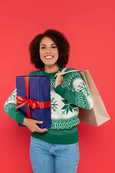 Mujer afroamericana feliz en suéter caliente sosteniendo bolsa de compras y caja de regalo azul aislado en rojo - foto de stock