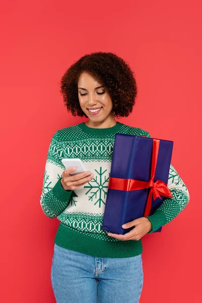 Mujer afroamericana feliz en suéter con patrón de invierno celebración de caja de regalo y teléfono móvil aislado en rojo - foto de stock