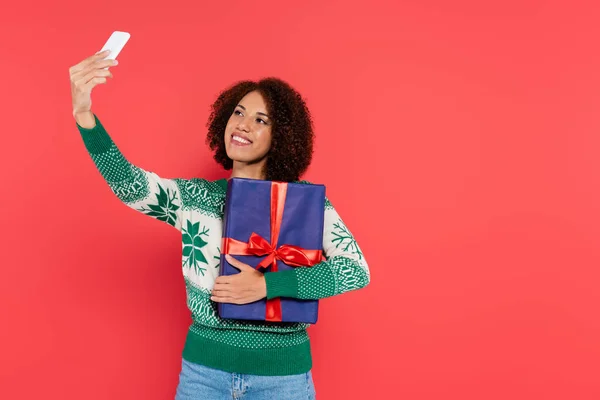 Felice donna africana americana in caldo maglione con motivo invernale prendendo selfie con scatola regalo blu isolato su rosso — Foto stock