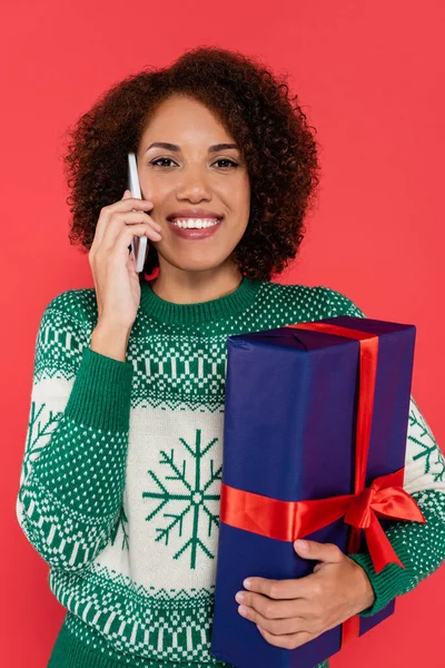 Alegre mujer afroamericana con caja de regalo azul hablando en el teléfono móvil aislado en rojo - foto de stock