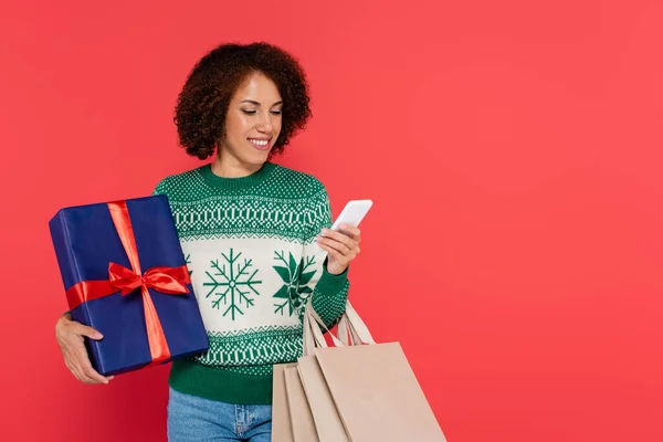 Sonriente mujer afroamericana con caja de regalo y bolsas de compras mirando teléfono inteligente aislado en rojo - foto de stock