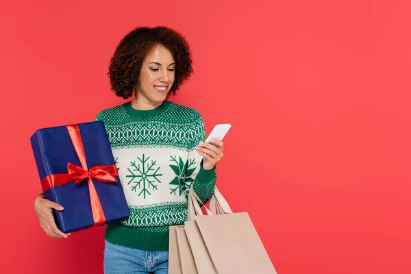 Femme afro-américaine souriante avec boîte cadeau et sacs à provisions regardant smartphone isolé sur rouge — Photo de stock