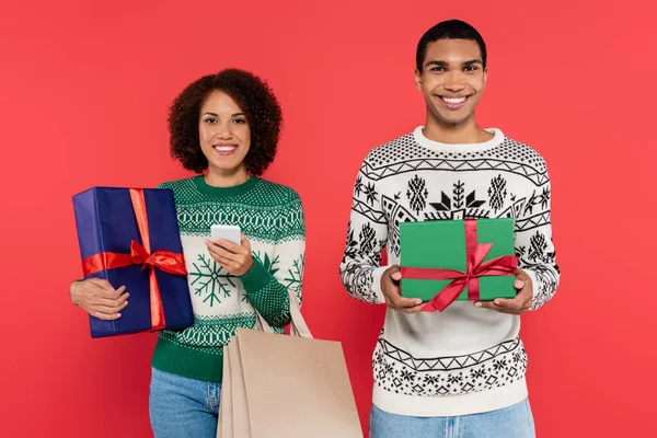 Donna afroamericana con smartphone in mano con borse della spesa e confezione regalo vicino all'uomo felice con presente isolato sul rosso — Foto stock