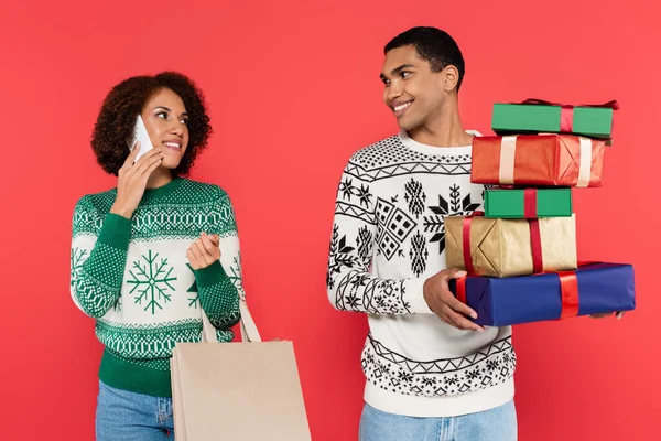 Afrikanisch-amerikanische Frau mit Einkaufstasche spricht auf Smartphone in der Nähe Mann mit bunten Geschenkboxen isoliert auf rot — Stockfoto