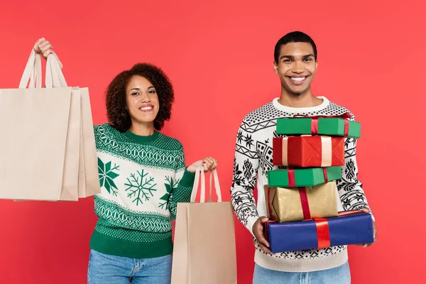 Pareja afroamericana joven con bolsas de compras y cajas de regalo mirando a la cámara aislada en rojo - foto de stock