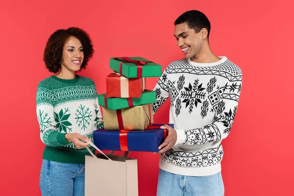 Happy african american man holding multicolored gift boxes near woman with shopping bag isolated on red — Stock Photo