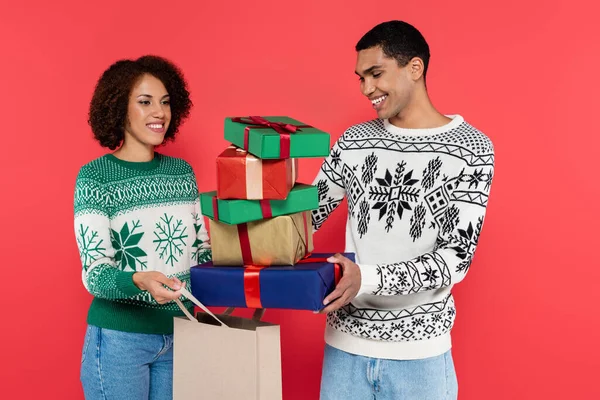 Felice uomo africano americano che tiene scatole regalo multicolore vicino alla donna con shopping bag isolato sul rosso — Foto stock