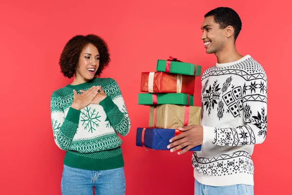 Erstaunt afrikanisch-amerikanische Frau berührt Brust neben Mann mit Stapel Weihnachtsgeschenke isoliert auf rot — Stockfoto