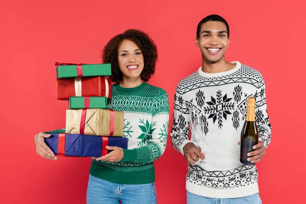 Sorrindo mulher afro-americana com caixas de presente multicoloridas perto do homem com champanhe isolado no vermelho — Fotografia de Stock