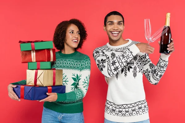 Joyeux homme afro-américain tenant des verres et bouteille de champagne près de petite amie avec des boîtes-cadeaux colorées isolées sur rouge — Photo de stock
