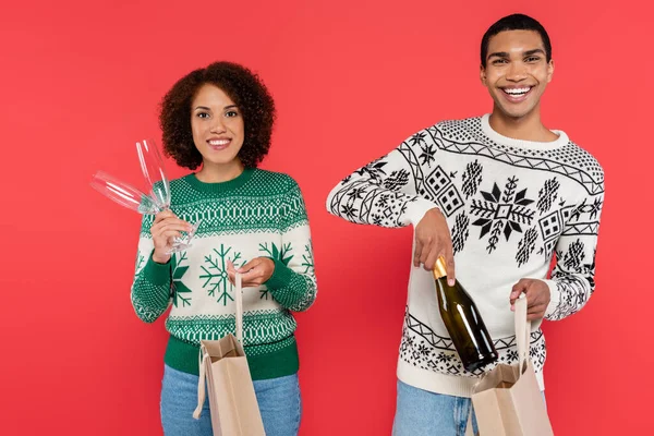 Felice coppia afro-americana con borse della spesa e champagne guardando la fotocamera isolata sul rosso — Foto stock