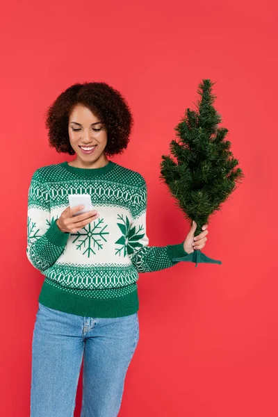 Sonriente mujer afroamericana en suéter caliente sosteniendo pequeño árbol de navidad y teléfono móvil aislado en rojo - foto de stock