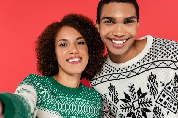 Jovem e feliz casal afro-americano em camisolas de inverno olhando para a câmera isolada no vermelho — Fotografia de Stock