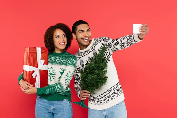 Homem americano africano alegre com pequena árvore de natal tomando selfie com namorada segurando presente isolado no vermelho — Fotografia de Stock