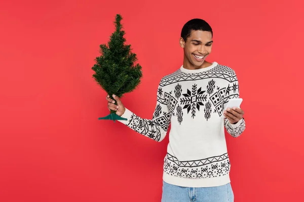 Homme afro-américain souriant en pull avec motif d'hiver tenant petit arbre de Noël et téléphone portable isolé sur rouge — Photo de stock