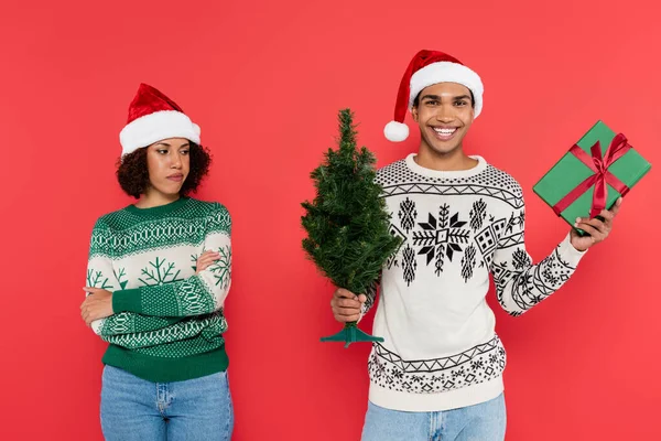 Gekränkte afrikanisch-amerikanische Frau mit Weihnachtsmütze steht mit verschränkten Armen neben glücklichem Mann mit Geschenk und Weihnachtsbaum auf rotem Grund — Stockfoto