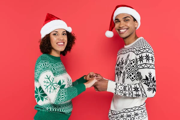 Glückliches afrikanisch-amerikanisches Paar in Winterpullovern und Weihnachtsmützen, Händchen haltend und isoliert auf rot in die Kamera blickend — Stockfoto