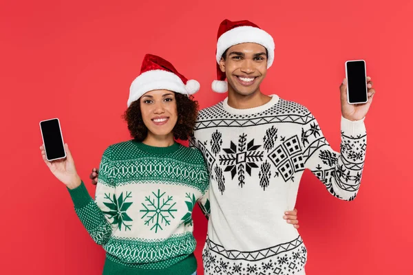 Glückliches afrikanisch-amerikanisches Paar in Weihnachtsmützen und warmen Pullovern, die Smartphones mit leerem Bildschirm zeigen, isoliert auf rot — Stockfoto