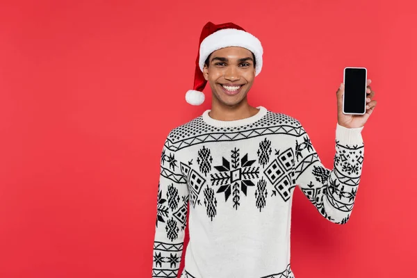 Homem americano africano feliz em chapéu de santa e suéter branco com padrão de inverno segurando smartphone com tela em branco isolado no vermelho — Fotografia de Stock