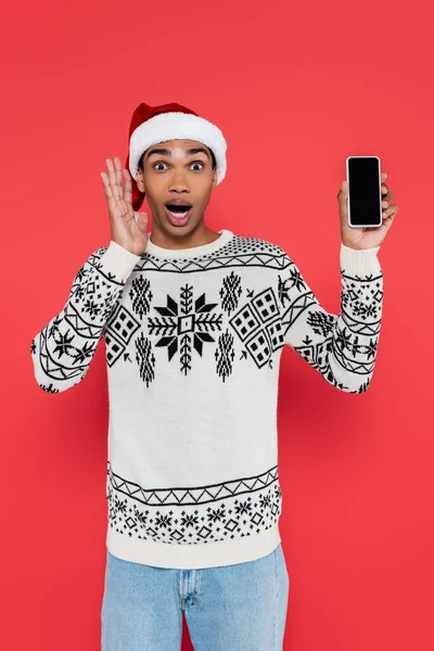 Espantado afro-americano homem em santa chapéu e suéter com ornamento de inverno segurando telefone celular com tela em branco isolado em vermelho — Fotografia de Stock