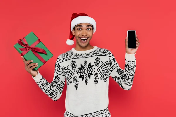 Hombre afroamericano emocionado en santa hat celebración de caja de regalo y teléfono celular con pantalla en blanco aislado en rojo - foto de stock