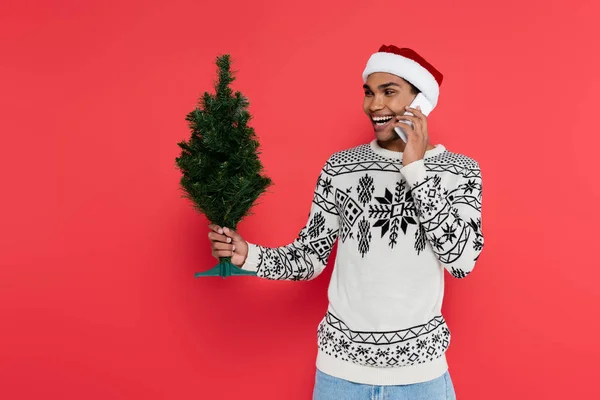 Homme afro-américain joyeux chapeau santa et pull chaud parler sur smartphone et tenant petit arbre de Noël isolé sur rouge — Photo de stock